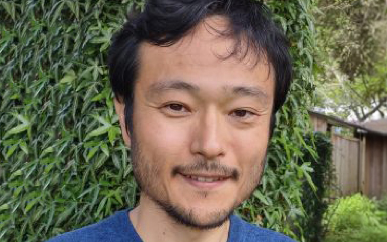 Koichi Haseyama, a man with short, slightly wavy black hair and facial hair smiles gently at the camera. He is wearing a blue T-shirt, and the background is an outdoor setting with green foliage and a wooden fence.