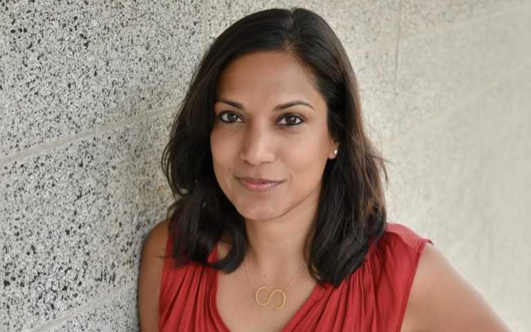 Surita Jhangiani, a woman with shoulder-length dark hair smiles softly while leaning against a light gray speckled wall. She is wearing a sleeveless red top and a necklace with a gold pendant shaped like the letter "S."